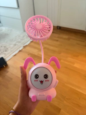 Söt rosa bordsfläkt med kaninöron - En liten, bärbar bordsfläkt i rosa plast med gulligt kaninansikte och kaninöron. Fläkten har ett flexibelt stativ så du kan vinkla den som du vill. Perfekt att ha på skrivbordet eller ta med i väskan för extra svalka.