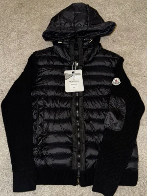 Svart Moncler pufferjacka med huva - Säljer en svart pufferjacka från Moncler i storlek M. Jackan har stickade ärmar, vadderad kropp och huva. Dragkedja framtill och ficka med dragkedja på ena ärmen. Klassisk Moncler-logga på vänster ärm. Perfekt för kalla vinterdagar. Den är Size M men jag skulle säga att den passar Size S bättre och om du är mellan 160-170 cm så skulle den sitta perfect. NFC-taggen fungerar och scannar utan problem.