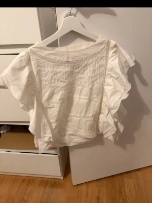 Vit blus med volangärmar - Säljer en söt vit blus med volangärmar och fina spetsdetaljer. Blusen har rund halsringning och dekorativa sömmar framtill.