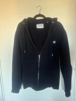 Marinblå zip hoodie med huva Les deux - En marinblå hoodie med dragkedja, huva och dragsnören. Klassisk passform med ribbade muddar vid ärmar och nederkant samt två fickor framtill. Liten patch på bröstet. Perfekt för en avslappnad och stilren look. Modell på bilden cirka 180cm alltså passar den för någon lite större än det