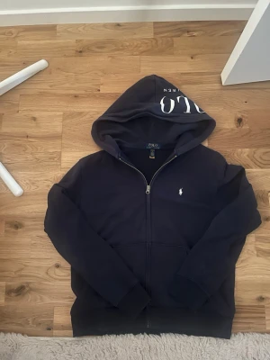 Marinblå hoodie från Polo Ralph Lauren - Snygg marinblå hoodie från Polo Ralph Lauren med dragkedja framtill och klassisk vit logga på bröstet. Stor huva med tryck och känguruficka. Perfekt för en chill och stilren look. Mjuk bomullskänsla och ribbade muddar vid ärmslut och nederkant.