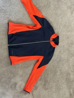 Marinblå och orange sportjacka Abacus - Snygg sportjacka från Abacus i marinblått och orange. Jackan har hel dragkedja framtill, hög krage och långa ärmar. Tillverkad i ett lätt och stretchigt material som passar perfekt för träning eller utomhusaktiviteter. Jag har använt till golf och det funkar perfekt 