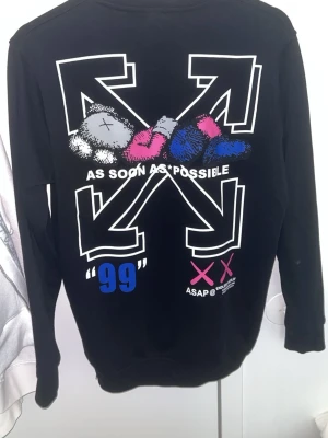 Svart Off-White hoodie med grafiskt tryck - Svart hoodie från Off-White med stort grafiskt tryck på ryggen i vitt, rosa, blått och grått. Texten 'AS SOON AS POSSIBLE' och '99' samt Off-White-logga och KAWS-inspirerade detaljer. Långärmad och i mjukt material, perfekt för en streetwear-look.