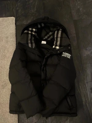Svart pufferjacka från Burberry - Säljer en svart pufferjacka från Burberry med huva och klassiskt rutigt innerfoder. Jackan har dragkedja framtill, två sidofickor och logotypen 'BURBERRY LONDON ENGLAND' på bröstet. Perfekt för kalla dagar och riktigt snygg streetstil priset kan diskuteras vid snabb affär