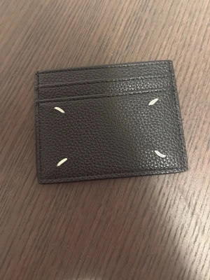 Maison margiela korthållare  - Stilrent svart kortfodral i grained läder med fyra vita sömdetaljer på framsidan. Fodralet har flera kortfack och en minimalistisk design som passar perfekt i fickan. Perfekt för dig som vill ha koll på dina kort utan att bära en stor plånbok.