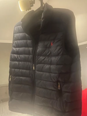 Svart dunväst från Polo Ralph Lauren - Snygg svart dunväst från Polo Ralph Lauren med klassisk röd logga på bröstet. Västen har hög krage, två dragkedjefickor och quiltad design. Perfekt för lager på lager och håller dig varm under kyliga dagar.