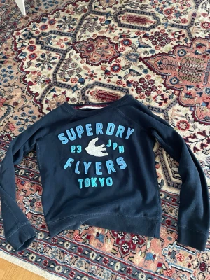 Mörkblå sweatshirt från Superdry - Mörkblå sweatshirt från Superdry med tryck i blått och vitt framtill där det står 'Superdry Flyers Tokyo' och en fågel. Tröjan har rund halsringning och långa ärmar. Perfekt för en avslappnad och sportig stil.