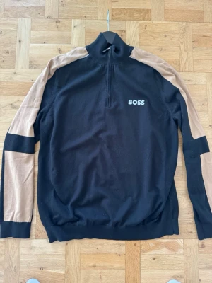 Svart och beige tröja från BOSS - Snygg svart tröja från BOSS med beige detaljer längs ärmarna och svart panel vid handlederna. Tröjan har halv dragkedja vid halsen och BOSS-logga på bröstet. Perfekt för en stilren och sportig look. använd fåtal gånger 