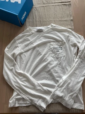 Acne Studios - Vit långärmad topp från Acne Studios med bröstficka där det står 'Acne Studios STOCKHOLM 1996' broderat i svart. Tillverkad i mjuk bomull och har en clean, minimalistisk look som passar till det mesta.