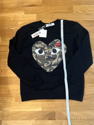 Svart sweatshirt med camo-hjärta Comme des garcons - Säljer en svart sweatshirt från Comme des Garçons Play med ett stort hjärta i camouflage-mönster och ögon på bröstet. Liten röd hjärtdetalj med öga på hjärtat. Långa ärmar, rund hals och ribbade muddar. Perfekt för dig som gillar streetwear och unika prints.