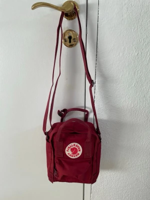 Vinröd Kånken Mini handväska - Säljer en vinröd Kånken Mini handväska från Fjällräven. Väskan har justerbar axelrem, två handtag upptill och klassisk rund logotyp framtill. Tillverkad i slitstarkt material och har en fyrkantig form med dragkedja upptill.