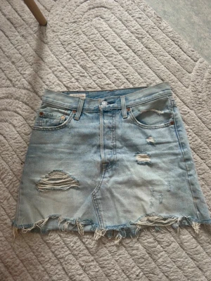 Ljusblå jeanskjol med slitningar - Säljer en kort ljusblå Levis jeanskjol med slitna detaljer och fransig nederkant. Kjolen har klassiska fickor fram och bak samt knapp och dragkedja i midjan. Perfekt för dig som gillar en avslappnad och trendig stil. Midjestorlek 26, skulle säga att den passar S.