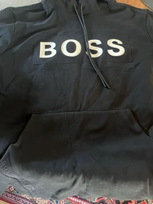 Svart hoodie från BOSS med logga - Snygg svart hoodie från BOSS med stor vit logga på bröstet och diskret BOSS-detalj på ärmen. Hoodien har huva med dragsko och en stor magficka. Tillverkad i mjukt material som känns skönt mot huden. Perfekt för en avslappnad och trendig look.