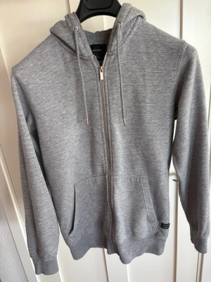 Grå zip hoodie med huva - Snygg grå hoodie med dragkedja framtill och två stora fickor. Tröjan har huva med dragsko och ribbade muddar vid ärmslut och nederkant. Perfekt för en avslappnad stil och enkel att matcha med jeans eller joggers.