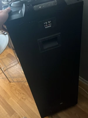 Large Black Speaker System - This image shows a large, black speaker system standing on a wooden floor. The top of the speaker has various control knobs and buttons, and there is a hand visible holding the side of the speaker. The front of the speaker has a handle and some input/output ports. The overall design is boxy and robust, suitable for events or parties.