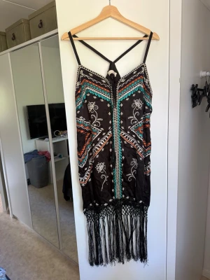 Svart boho klänning med fransar - Unik svart klänning med tunna axelband och färgglada boho-mönster i turkos, orange och vitt. Klänningen har långa fransar nedtill och ett korsat band i ryggen. Perfekt för sommarens festivaler eller strandhäng. Från märket Odd Molly i storlek 3 som enligt deras storlekguide motsvarar en storlek L. Mycket fint skick, jag hittar inga synliga anmärkningar eller defekter. 