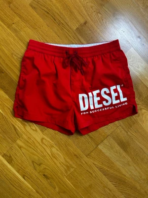 Röda badbyxor från Diesel - Snygga röda badbyxor från Diesel med stort vitt logotryck på ena benet. De har resår och dragsko i midjan samt innerbyxa i mesh. Perfekta för stranden eller poolen och riktigt trendiga med sin starka färg och tydliga branding.
