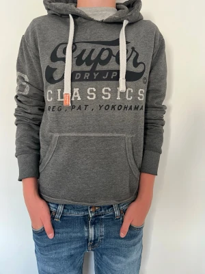 Grå hoodie från Superdry - Säljer en grå hoodie från Superdry med stor logga och text på bröstet. Hoodien har vita snören, känguruficka och ribbade muddar. Perfekt för dig som gillar en avslappnad och sportig stil.