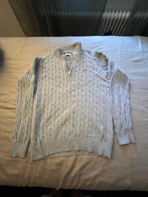 Ljusgrå kabelstickad tröja Gant - Kabelstickad ljusgrå tröja från Gant med halv dragkedja och ståkrage. Tröjan har ribbade muddar och nederkant samt Gant-logga broderad på bröstet. Perfekt för dig som gillar klassisk och stilren look.