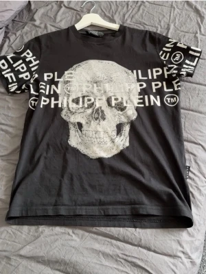 Svart t-shirt med dödskalletryck - Svart t-shirt med stort dödskalletryck framtill och texten 'PHILIPP PLEIN' i vitt över bröstet och på ärmarna. T-shirten har rund hals och korta ärmar. Materialet känns mjukt och skönt, perfekt för en edgy streetstyle. Ni får både tjock tröjan och t shirt t shirt storlek s tjocktröjan storlek m