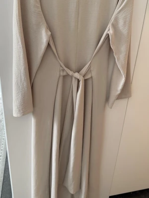 Långärmad maxiklänning med knytband  - En stilren beige långklänning med rund halsringning och trekvartsärmar. Klänningen har en rak passform och är tillverkad i ett mjukt, lätt material som ger ett elegant fall.
