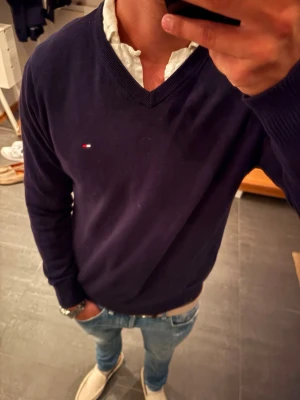 Mörkblå V-ringad tröja Tommy Hilfiger - Klassisk mörkblå V-ringad tröja från Tommy Hilfiger med liten broderad logga på bröstet. Tröjan har långa ärmar och ribbade muddar vid ärmslut och nederkant. Perfekt att bära över en skjorta för en clean och stilren look. M passar S också. Modellen är 180 cm och 65 kg