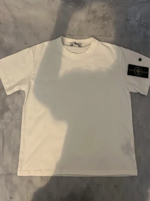 Vit t-shirt med patch på ärmen - Vit t-shirt i klassisk modell med rund hals och korta ärmar. På vänster ärm sitter en svart patch med logga och knappdetalj. Perfekt basplagg med en clean look och snygg detalj på ärmen.