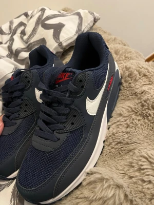 Nike Air Max 90 marinblå sneakers - Snygga Nike Air Max 90 sneakers i marinblått med vita detaljer och röda inslag. Ovandelen är i mesh och syntet, med den klassiska vita swoosh-loggan på sidan och synlig Air Max-dämpning i sulan. Perfekta för dig som gillar sportig och stilren look.