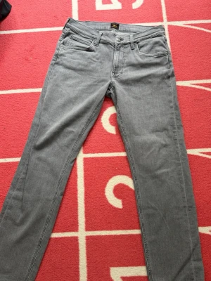 Grå raka jeans från Lee - Snygga grå jeans från Lee med rak passform och klassisk femficksdesign. Jeansen har normal midja och är tillverkade i ett mjukt denimtyg som ger en avslappnad look. Perfekta för dig som gillar stilrena och tidlösa plagg. Använd 2ggr så perfekt skick!