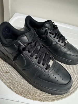 Svarta Nike Air Force 1 sneakers - Klassiska helsvarta Nike Air Force 1 sneakers i läder med svart sula och svarta skosnören. Ikonisk design med perforerad tå och metallplatta på snörningen. Perfekta för en clean och stilren look.