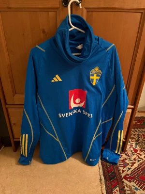 Blå svensk landslagströja Adidas - Blå långärmad landslagströja från Adidas med Sveriges fotbollsförbunds logga och Svenska Spel-tryck på bröstet och ryggen. Tröjan har gula detaljer och ränder på ärmarna samt hög krage. Perfekt för dig som vill stötta Sverige på match!