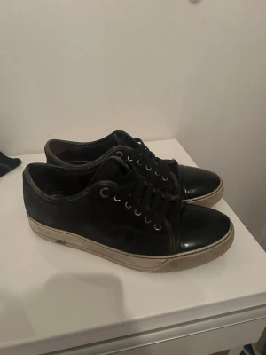 Lanvins sneakers med gummisula - Lanvin sneakers i svart läder, originalpåse ingår. Köpta begagnade och använda varsamt i cirka ett år. Skosnöre och sula är skadat. Kom med frågor angående skadorna. Nypris cirka 5 000 kr. Påse till skorna ingår. Prisförslag fungerar vid snabb affär.