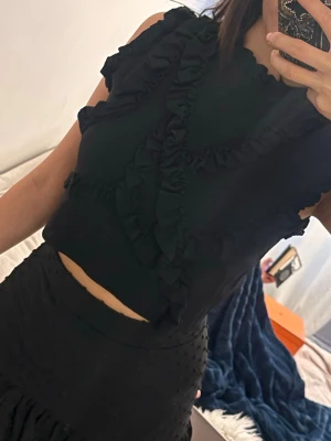 Svart croppad volangtop - Säljer en svart croppad topp med volangdetaljer framtill och längs ärmhålen. Toppen är ärmlös och har en rund halsringning. Perfekt för dig som gillar en trendig och feminin stil. Storlek m men passar xs 