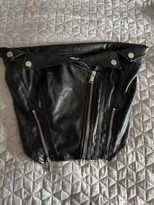 Svart skinn korsett-topp med dragkedjor - Cool svart korsett-topp i skinnimitation med dragkedjor och silvriga knappar framtill. Toppen är axelbandslös och har en edgy bikerstil, perfekt för att sticka ut. Snygg detalj med flera dragkedjor och markerad midja.