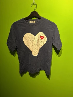 Blå Comme des Garçons Play t-shirt - Mörkblå t-shirt från Comme des Garçons Play med stort vitt hjärta och röd hjärtdetalj på bröstet. Klassisk rund hals och korta ärmar. Perfekt för dig som gillar streetwear och vill sticka ut med en ikonisk design. Lite slitage på designen som syns på bilder😊