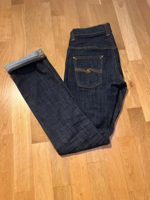 Mörkblå jeans från Nudie Jeans - Snygga mörkblå jeans från Nudie Jeans med klassisk femficksdesign och kontrastsömmar. Jeansen har en rak passform och är tillverkade i kraftig denim. Bakfickan har Nudie Jeans ikoniska broderi och läderpatch i midjan.