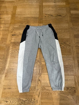 Nike grå mjukisbyxor med färgblock - Snygga mjukisbyxor från Nike i grått med vita och svarta färgblock på sidorna. Byxorna har resår i midjan med dragsko och muddar vid bensluten. Perfekta för chill dagar eller träning. Klassisk Nike-logga på baksidan.