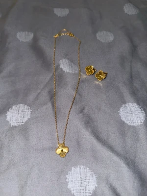 Guldfärgat halsband med blomma - Säljer ett tunt guldfärgat halsband med en söt blomformad berlock. Kedjan är finlänkad och har ett klassiskt lås. Matchande örhängen i samma blommiga design ingår. Perfekt accessoar för att lyfta din outfit med en touch av guld och blommor.