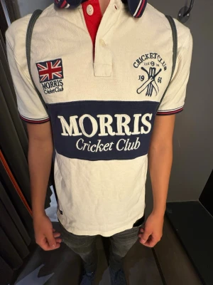 Vit piké Morris Cricket Club - Vit pikétröja från Morris Cricket Club med blåa och röda detaljer, broderad logga och brittisk flagga på bröstet. Stora tryckta bokstäver över magen och klassisk krage med knappar. Perfekt för en sportig och stilren look. Är L men passar 172-180 jätte bra. Är 177cm 70kg och sitter perfekt 