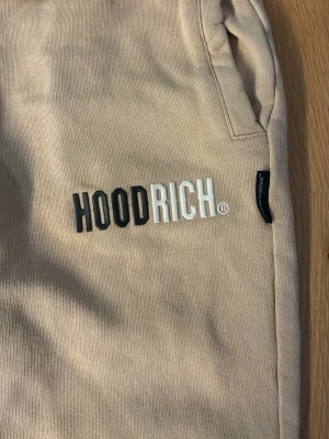 Beige mjukisbyxor från Hoodrich - Snygga beige mjukisbyxor från Hoodrich med broderad logga på benet. Byxorna har resår i midjan med dragsko, fickor på sidorna och muddar vid bensluten. Perfekta för en avslappnad och trendig stil.