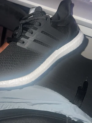 Svarta sneakers ultra boost  - Snygga svarta sneakers med stickad ovandel och vit, dämpad sula. Skorna har svarta snören och detaljer i plast längs sidorna. Perfekta för en sportig och stilren look. Låg modell som passar till många outfits.