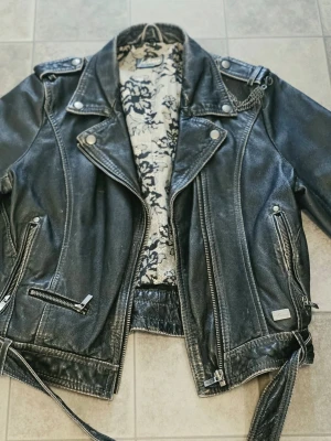 Jofama by Kenza skinnjacka – Vintage / Patina – Bikerjacka strl 40 - Säljer en riktigt snygg bikerjacka i äkta skinn från Jofama by Kenza. ​Jackan har en fantastisk vintage-look med naturlig patina och slitningar i lädret som ger den en cool och unik karaktär. Snyggt mönstrat innerfoder i spetsmönster, detaljer som kedja på axeln, nitar baktill, bälte samt dragkedjor på ärmarna. ​Märke: Jofama (Kenza collection) ​Modell: Bikerjacka / Läderjacka ​Storlek: 40 ​Material: Äkta läder / skinn ​