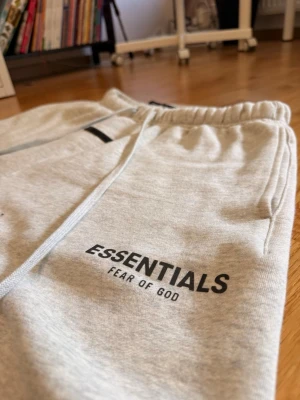 Light oat essentials shorts - Snygga light oat från Essentials Fear of God med elastisk midja och långa snören framtill. Mjuk bomullskänsla och logga tryckt på vänster ben. Perfekta för chill dagar eller streetwear-stil.
