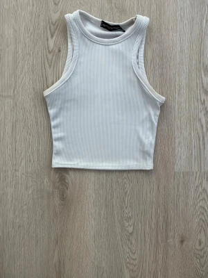 Vit ribbad ärmlös linnetopp - Snygg vit ribbad linnetopp med hög hals och ärmlös design. Perfekt att matcha med jeans eller kjol för en clean och fräsch look. Croppad modell som ger en trendig siluett.