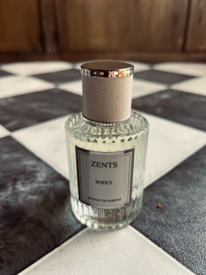 Zents Wifey Extrait de Parfum - Elegant parfymflaska från Zents med doften Wifey. Flaskan är genomskinlig med räfflad glasdesign och har en vit etikett. Locket är vitt med silverdetaljer och har en lyxig känsla. Perfekt för dig som vill ha en exklusiv och stilren parfym i samlingen. Bara testad