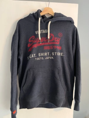 Mörkblå Superdry hoodie med tryck - Mörkblå hoodie från Superdry med rött och vitt tryck på bröstet. Klassisk känguruficka framtill, vita dragsnören i huvan och ribbade muddar. Perfekt för en avslappnad och cool stil.