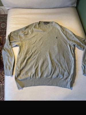 Grå stickad tröja med V-ringning - Snygg grå stickad tröja från Polo Ralph Lauren med klassisk V-ringning och den ikoniska gröna loggan broderad på bröstet. Tröjan har långa ärmar och ribbade muddar vid ärmslut och nederkant. Perfekt för lager på lager och stilren vardagslook. 100% pima cotton