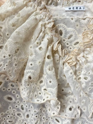 Vit broderad blus från Wera - Romantisk vit blus från Wera med broderade hålmönster och volangdetaljer vid halsen. Blusen är ärmlös och har en luftig, lätt transparent känsla. Perfekt för dig som gillar en feminin och trendig stil.