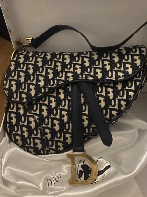 Dior Saddle Bag i blått canvas -  Dior Saddle Bag i blå och beige canvas med det klassiska Dior-monogrammet över hela väskan. Väskan har guldfärgade metalldetaljer, bland annat ett stort D-spänne framtill och C-spännen på axelremmen. Snygg och trendig form som sticker ut.