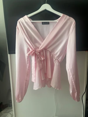 Ljusrosa omlottblus i satin - Supersöt ljusrosa omlottblus från Missay i glansigt satinmaterial. Blusen har långa ärmar, knytband i midjan och en v-ringad omlottfront som ger en feminin siluett. Perfekt att styla med jeans eller kjol för en trendig look.